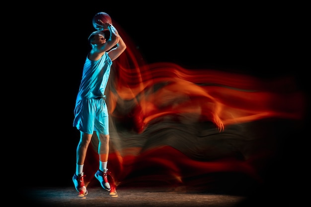 Männlicher Basketballspieler, der Basketball einzeln über dunkler Studiowand in gemischtem Licht spielt.