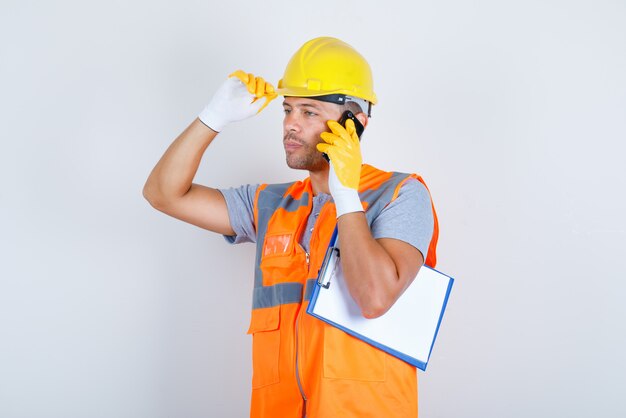 Männlicher Baumeister, der am Telefon mit Hand auf Helm in Uniform, Handschuhen, Vorderansicht spricht.