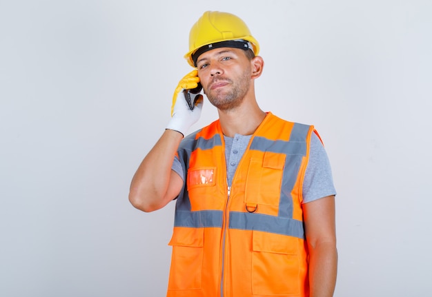 Männlicher Baumeister, der auf Handy in Uniform, Helm, Handschuhen, Vorderansicht spricht.