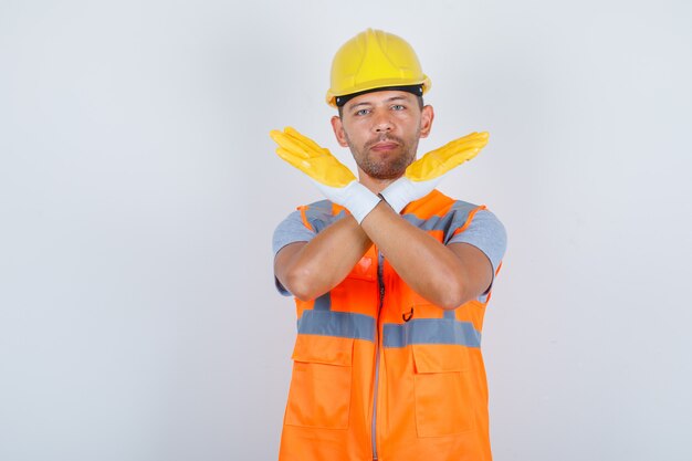 Männlicher Baumeister, der kein Zeichen mit verschränkten Armen in Uniform, Helm, Handschuhen, Vorderansicht gestikuliert.