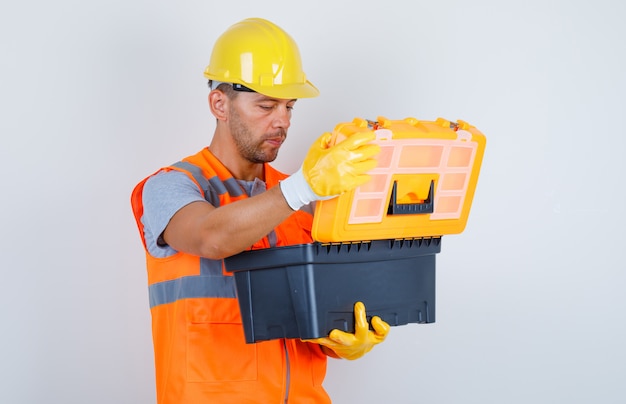 Männlicher Baumeister, der Kunststoff-Werkzeugkasten in Uniform, Helm, Handschuhen, Vorderansicht öffnet.