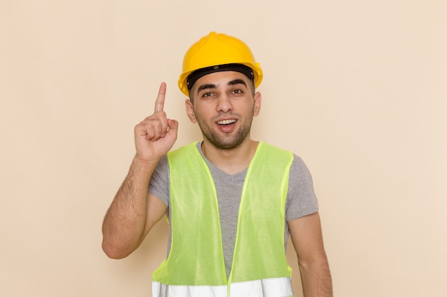 Männlicher Baumeister der Vorderansicht im gelben Helm, der mit erhobenem Finger auf dem hellen Hintergrund aufwirft