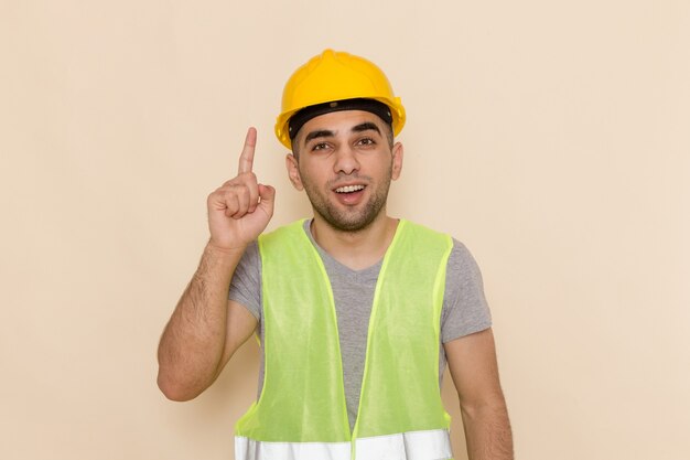 Männlicher Baumeister der Vorderansicht im gelben Helm, der mit erhobenem Finger auf dem hellen Hintergrund aufwirft