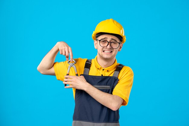 Männlicher Baumeister der Vorderansicht in der Uniform und im Helm, die seinen Finger mit einer Zange auf Blau schneiden