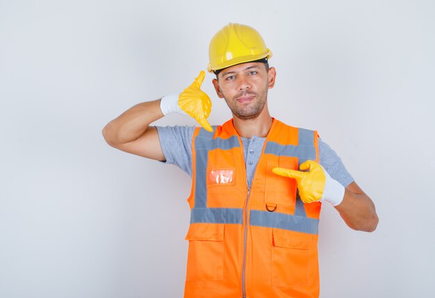 Männlicher Baumeister in Uniform, Helm, Handschuhen, die mich anrufen oder Kontaktgeste zeigen und selbstbewusst aussehen, Vorderansicht.