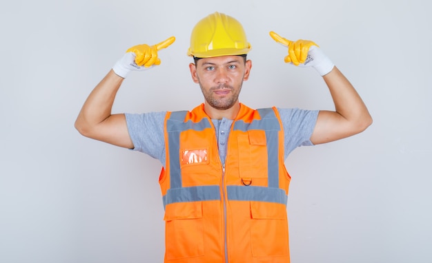 Männlicher Baumeister in Uniform zeigt Finger auf Sicherheitshelm und schaut selbstbewusst, Vorderansicht.
