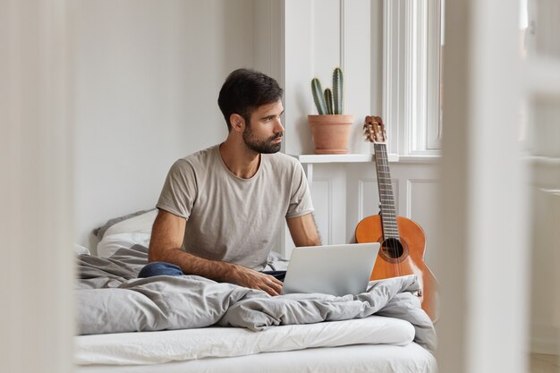 Männlicher Blogger hat nachdenklichen Ausdruck, genießt Freizeit mit Laptop, sitzt auf bequemem Bett