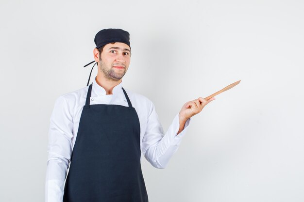 Männlicher Chef, der hölzernen Spatel in Uniform, Schürze hält und fröhlich schaut. Vorderansicht.