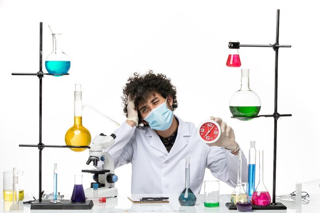 Männlicher Chemiker der Vorderansicht im medizinischen Anzug und mit der Maske, die rote Uhren auf weißem Schreibtisch hält