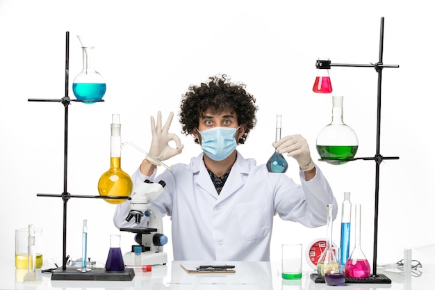 Männlicher Chemiker der Vorderansicht im weißen medizinischen Anzug und mit Maske, die blaue Lösung auf hellem weißem Raum hält