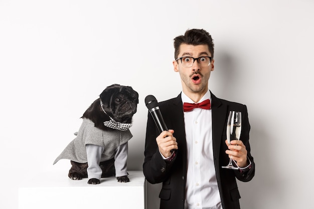 Männlicher Entertainer, der ein Glas Champagner anhebt, dem süßen schwarzen Hund ein Mikrofon gibt, der auf weißem Hintergrund steht