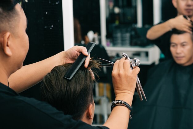 Männlicher Friseur, der das Haar des Kunden mit Baut. Und Scheren vor Spiegel schneidet
