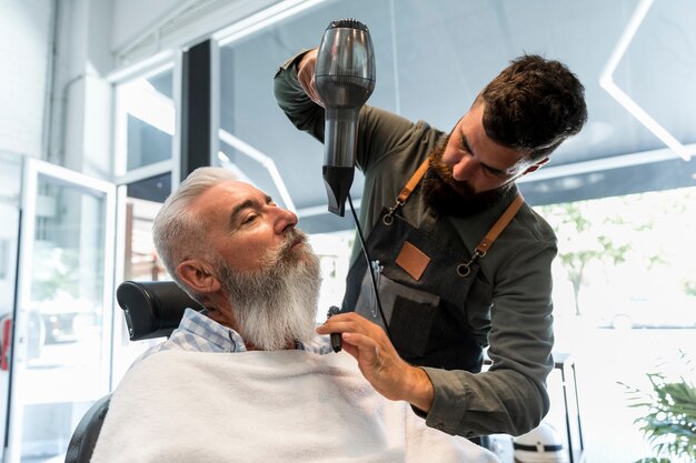 Männlicher Friseur, der Trockner für Bart des älteren Kunden verwendet