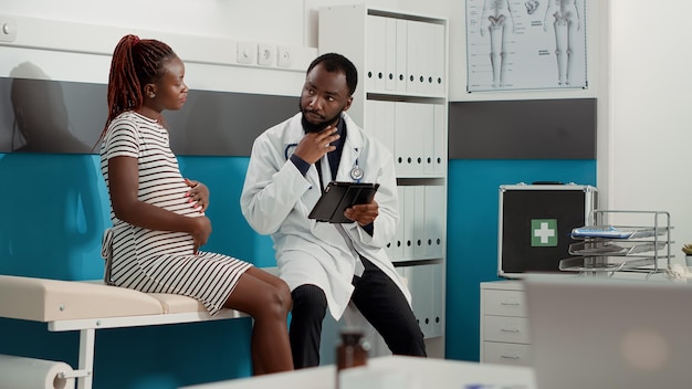 Männlicher Geburtshelfer, der sich beim Kontrollbesuch Notizen auf einem digitalen Tablet macht und mit einer Patientin mit Schwangerschaftsbauch in der Arztpraxis spricht. Arzt konsultiert werdende Frau mit Babybauch, um Ratschläge zu geben.