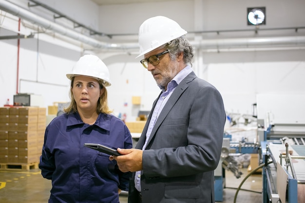 Männlicher Ingenieur und weiblicher Fabrikarbeiter in Schutzhelmen, die auf Pflanzenboden stehen und sprechen, Mann, der Tablette verwendet