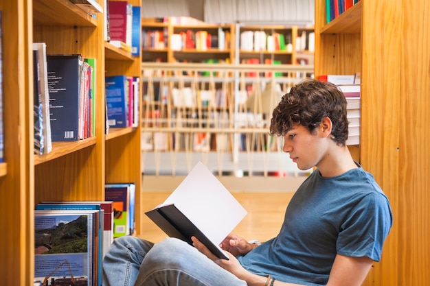 Kostenloses Foto männlicher jugendlicher, der auf bücherschrank und lesung sich lehnt