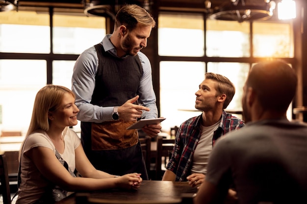 Männlicher Kellner, der Touchpad verwendet, während er Kunden in einem Café bedient und ihre Bestellungen entgegennimmt