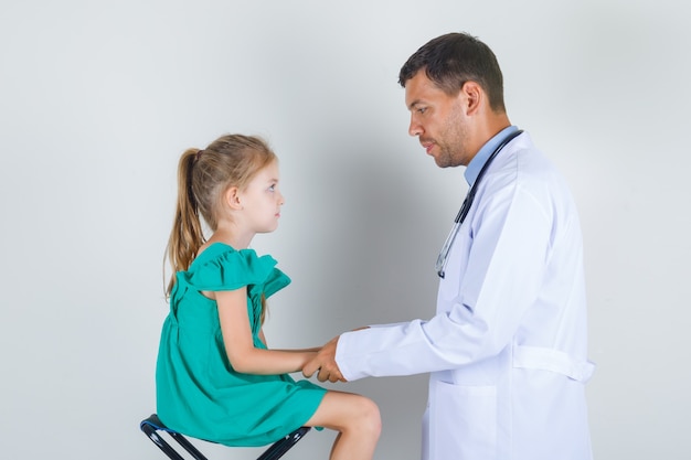 Männlicher Kinderarzt, der kleines Mädchen in weißen Kleidern untersucht und vorsichtig schaut. Vorderansicht.