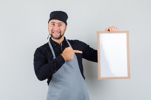 Männlicher Koch, der auf weiße Tafel im Hemd, in der Schürze zeigt und glücklich schaut, Vorderansicht.