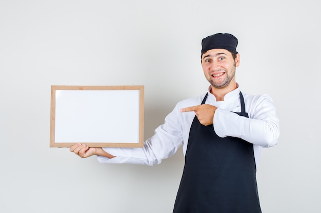 Männlicher Koch, der auf weiße Tafel in Uniform, Schürze zeigt und fröhlich schaut. Vorderansicht.
