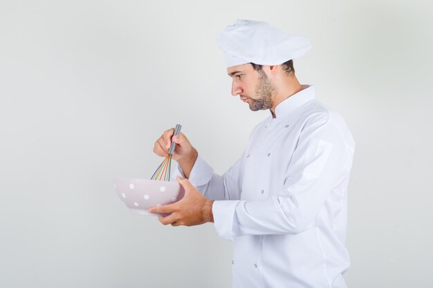 Männlicher Koch, der Bestandteile mit Schneebesen in Schüssel in weißer Uniform mischt und beschäftigt schaut