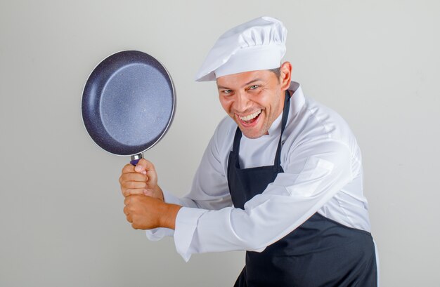 Männlicher Koch, der Bratpfanne hält, während Spaß in Hut, Schürze und Uniform hat und amüsant aussieht