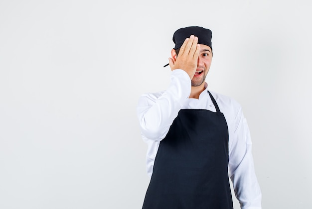 Männlicher Koch, der ein Auge mit Hand in Uniform, Schürze bedeckt und fröhlich, Vorderansicht schaut.