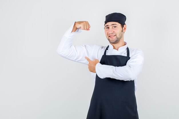 Männlicher Koch, der Finger auf seinen Muskel in Uniform, Schürze zeigt und selbstbewusst aussieht. Vorderansicht.