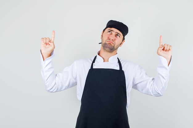 Männlicher Koch, der Finger in Uniform, Schürze zeigt und düster, Vorderansicht schaut.