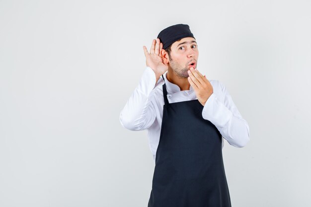Männlicher Koch, der Hand hinter Ohr hält, um in Uniform, Schürze zu hören und überrascht auszusehen. Vorderansicht.