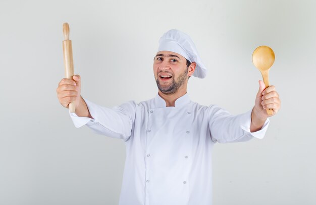 Männlicher Koch, der Holzlöffel und Nudelholz in weißer Uniform hält und fröhlich schaut