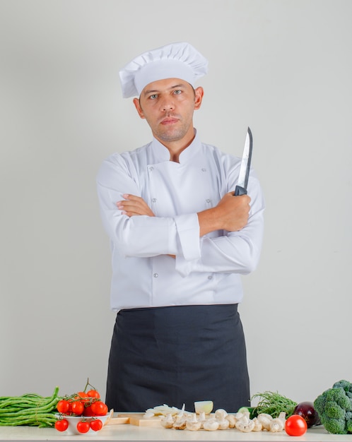 Männlicher Koch, der Messer mit verschränkten Armen in der Küche in Uniform, Hut und Schürze hält