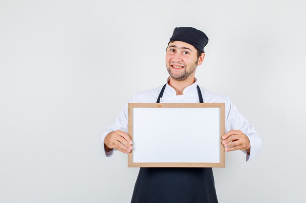 Männlicher Koch, der weiße Tafel in Uniform, Schürze hält und fröhlich schaut. Vorderansicht.