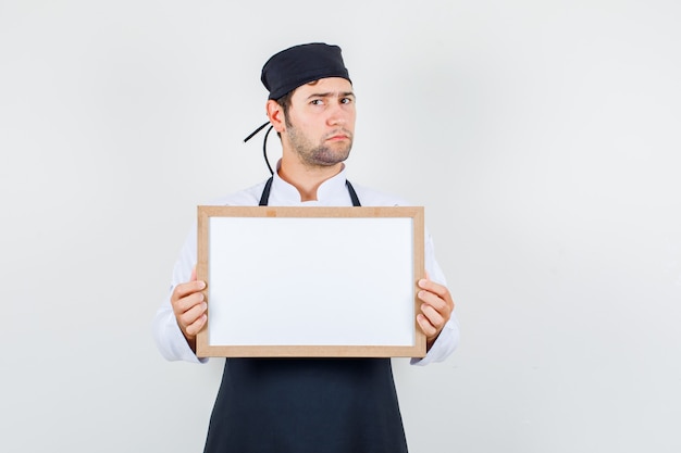 Männlicher Koch, der weiße Tafel in Uniform, Schürze hält und verärgert schaut. Vorderansicht.