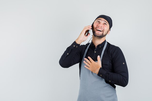 Männlicher Koch im Hemd, Schürze, die auf Handy spricht und fröhlich, Vorderansicht schaut.