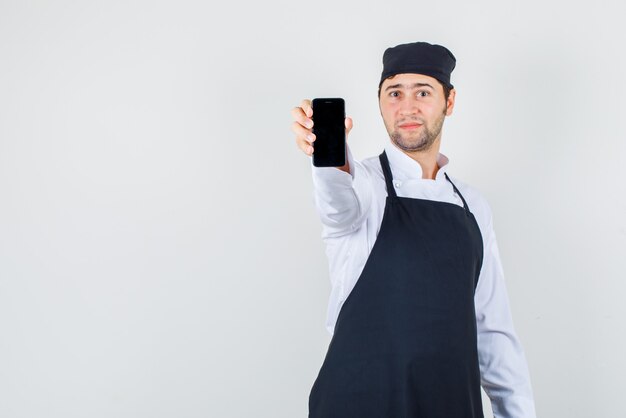 Männlicher Koch in Uniform, Schürze, die Handy zeigt und fröhlich schaut, Vorderansicht.