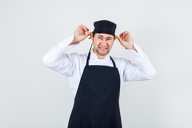 Kostenloses Foto männlicher koch in uniform, schürze, die ohren zieht, vorderansicht.