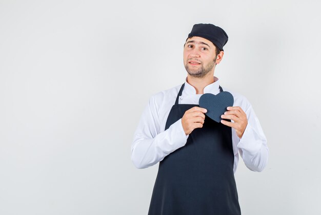 Männlicher Koch in Uniform, Schürze, die schwarze Geschenkbox hält, Vorderansicht.