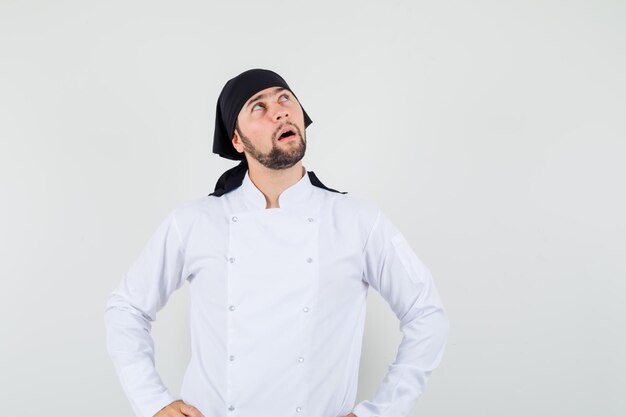 Männlicher Koch in weißer Uniform, der mit den Händen auf der Taille nach oben schaut und nachdenklich aussieht, Vorderansicht.