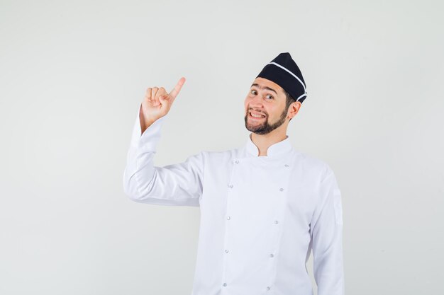 Männlicher Koch in weißer Uniform, der nach oben zeigt und froh aussieht, Vorderansicht.