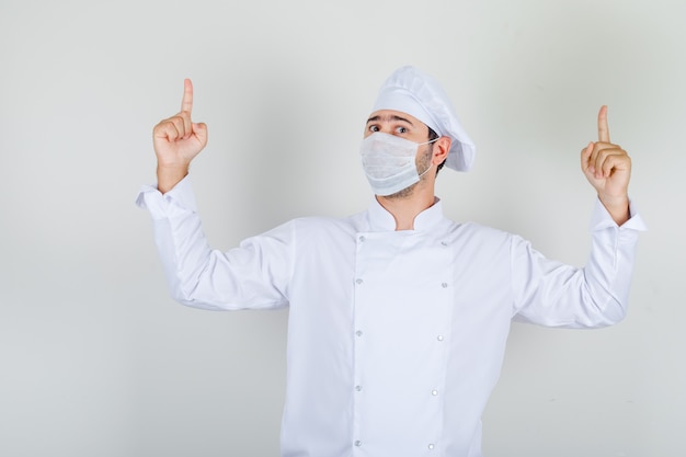 Männlicher Koch in weißer Uniform, medizinische Maske, die Finger nach oben zeigt und vorsichtig schaut