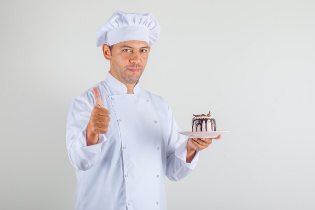 Männlicher Koch kocht, der Kuchen hält und Daumen in Hut und Uniform zeigt