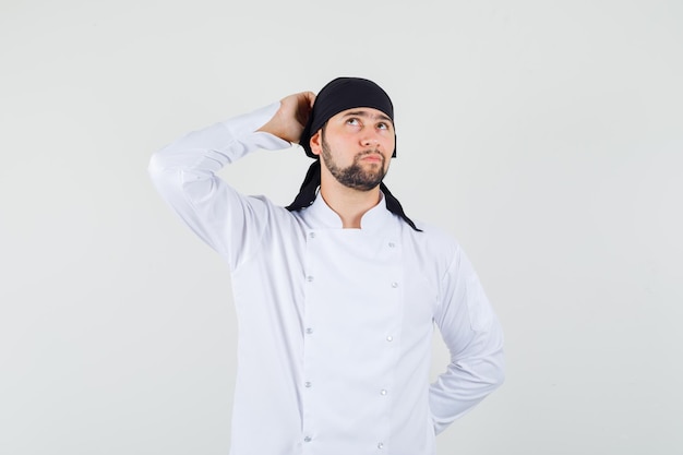 Männlicher Koch kratzt sich am Kopf, während er in weißer Uniform nach oben schaut und nachdenklich aussieht, Vorderansicht.