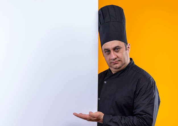 männlicher Koch mittleren Alters in Kochuniform, die weiße Wand auf gelber Wand mit Kopienraum hält