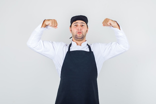 Männlicher Koch zeigt seine Muskeln in Uniform, Schürze und sieht selbstbewusst aus. Vorderansicht.