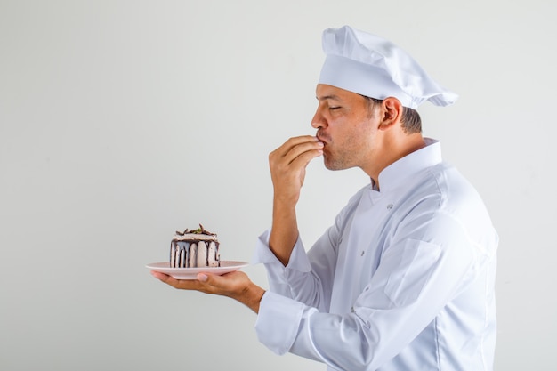 Männlicher kochkoch, der kuchen hält und leckere geste in hut und uniform macht