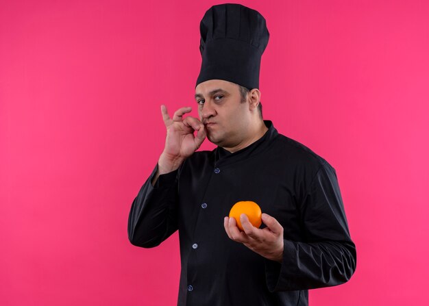 Männlicher Kochkoch, der schwarze Uniform und Kochhut hält, der orange Frucht zeigt Zeichen für köstliches Stehen über rosa Hintergrund