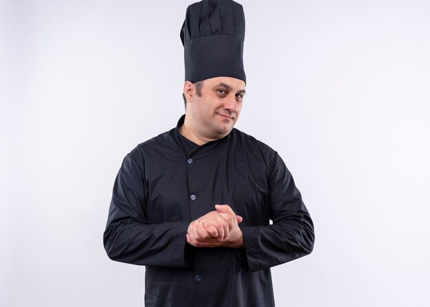 Männlicher Kochkoch, der schwarze Uniform und Kochhut trägt, die Kamera lächelnd reibende Hände steht über weißem Hintergrund