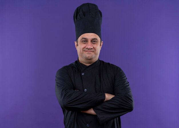 Männlicher Kochkoch, der schwarze Uniform und Kochhut trägt Kamera mit sicherem Lächeln auf Gesicht mit gekreuzten Händen auf Brust, die über lila Hintergrund steht