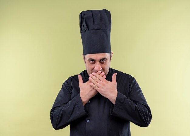 Männlicher Kochkoch, der schwarze Uniform und Kochhut trägt und die Kamera betrachtet, die schlau den Mund mit den Händen bedeckt, die über grünem Hintergrund stehen
